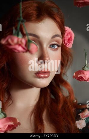 Red-haired woman with striking blue eyes looks away, surrounded by delicate pink roses Stock Photo