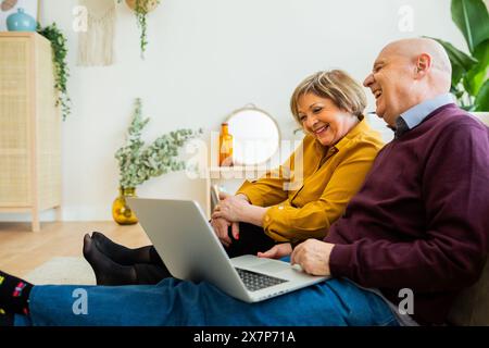 Senior couple having a video call on a laptop, smiling at the screen. Senior couple in a living room, connecting with loved ones online. Stock Photo