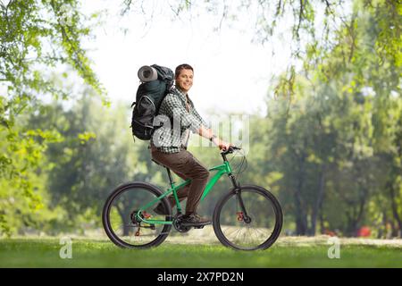 Full length profile shot of a young man with a backpack riding a bicycle in a park and looking at camera Stock Photo