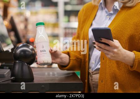Close up of unrecognizable woman scanning milk bottle while using self checkout service grocery shopping and using smartphon Stock Photo