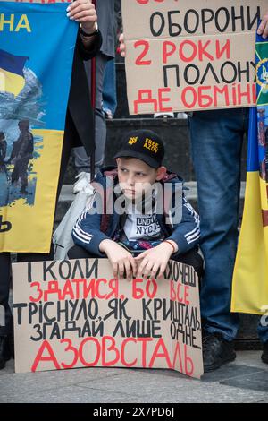 May 18, 2024, Lviv, Ukraine: A little boy with a banner sits on the curb during the protest in the center of Lviv. Relatives and friends of captured defenders of Mariupol, holding banners and flags, participated in the ''Don't be silent. Capture kills. Two years of captivity'' protest in Lviv. The event, organized by the Association of Families of Defenders of Azovstal, commemorated the anniversary of the captivity of Ukrainian defenders from the Azovstal plant. On May 20, 2022, these defenders left the plant and were captured by the Russians. Over 2,000 Ukrainian soldiers remain in captivity Stock Photo