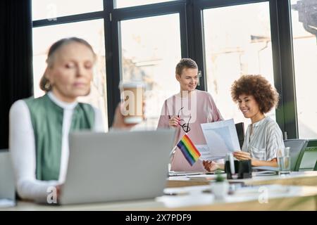 Blurred mature woman works on a laptop at a table, with her focused diverse colleagues on backdrop. Stock Photo
