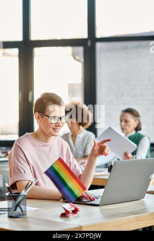 Short haired woman engrossed in work, with a laptop in front of her, with her diverse colleagues on backdrop, pride flag. Stock Photo