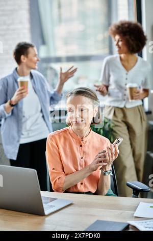 Merry businesswoman talking on phone while using laptop, with her diverse colleagues on backdrop. Stock Photo