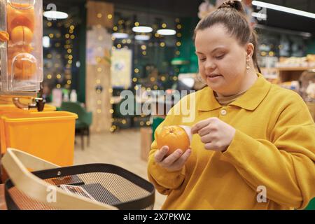 Portrait of cheerful young woman with Down syndrome buying fresh oranges in fruit section of supermarket and wearing yellow Stock Photo