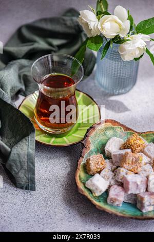 Assorted traditional Turkish delights on a ceramic plate beside a glass of black tea, with a vase of white roses in the background Stock Photo
