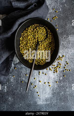 A rustic setting displays raw green mung beans scattered around and within a dark bowl, accompanied by a metal spoon on a textured, grey surface Stock Photo