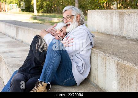 A senior couple shares a joyful hug while sitting on a concrete ledge, radiating happiness and companionship in a serene park setting Stock Photo