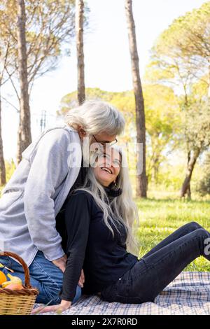 An elderly couple shares a laugh while enjoying a romantic picnic on a sunny day in a lush park, surrounded by tall trees and nature Stock Photo