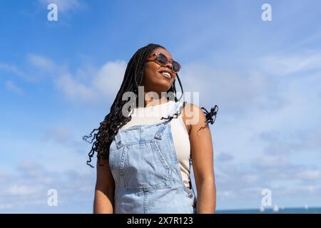 A joyful woman in a denim jumpsuit smiles under a clear blue sky, embodying the carefree spirit of summer in Barcelona Stock Photo
