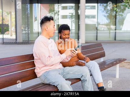 An Asian male and African female sit closely, smiling and focused on a smartphone screen together, conveying a sense of connection and technology use Stock Photo
