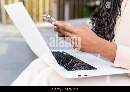 Close-up of an African American businesswoman using her smartphone while typing on a laptop, focusing on multitasking with technology outdoors Stock Photo