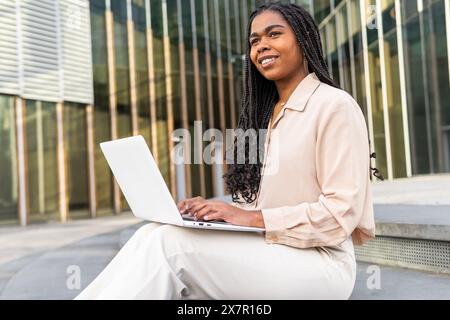 A smiling woman enjoys working on her laptop outdoors in a modern urban setting during a sunny day in Barcelona Stock Photo