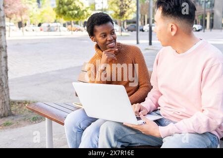 Two students, a Black woman and an Asian man, engage in discussion while looking at each other while working in a laptop together, seated on a park be Stock Photo