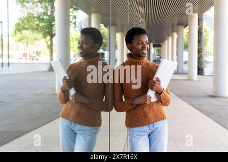 A young African female student smiling, holding a laptop, and standing near a reflective glass wall. Stock Photo