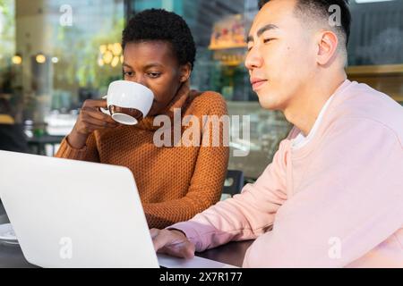 A young African female student sipping coffee while looking at a laptop screen next to an Asian male student in a cafe. Stock Photo