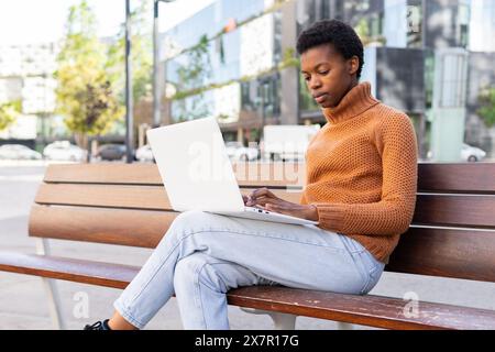A focused African American female student works on her white laptop while sitting on a bench outdoors, deeply engrossed in her task. Stock Photo