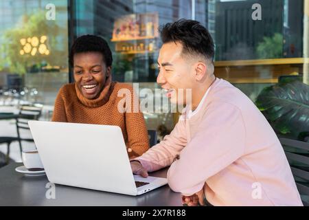 Two young students of different ethnicities enjoy a cheerful moment together while looking at a laptop screen in a cafe setting. Stock Photo