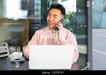 Smiling Asian man in casual attire having a conversation on his smartphone while working on a laptop in a cafe setting, with a cup of coffee on the ta Stock Photo