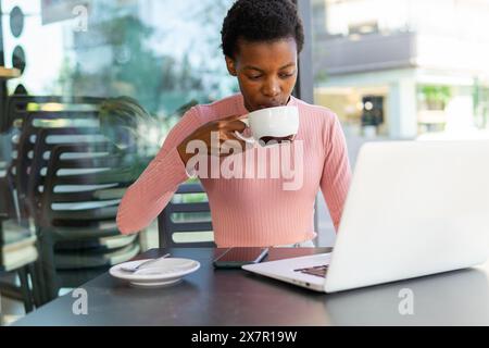 An African woman is focused on her laptop screen while sipping coffee at a cafe, reflecting productivity and a casual work environment. Stock Photo