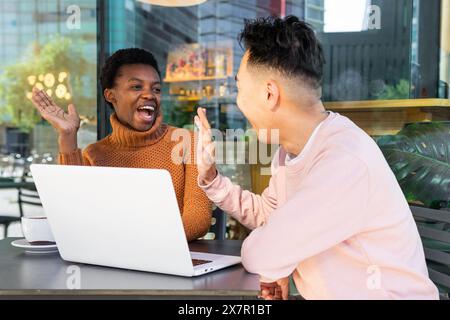 A joyful African female student and an Asian male student engage in animated conversation at an outdoor cafe with a laptop on the table. Stock Photo