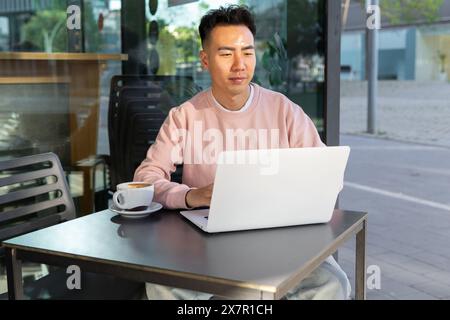 An Asian man is looking at his laptop screen, engaged in work at an outdoor cafe, with a coffee cup in sight. Stock Photo