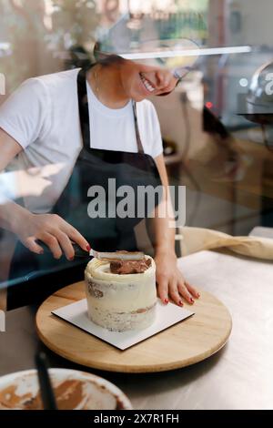 A smiling pastry chef, viewed through a protective glass, skillfully spreads chocolate frosting on a multi-layered cake inside a bright, modern bakery Stock Photo