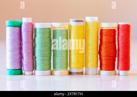 A line of vibrant sewing thread spools organized by color, including purple, green, yellow, and red, on a light table surface against a plain backgrou Stock Photo