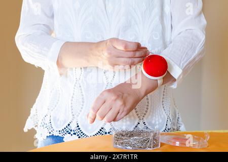 A close-up of a seamstress threading a needle with a pin cushion on her wrist, next to a box of pins Stock Photo