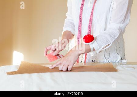 A tailor with a measuring tape around her neck is marking a pattern on brown fabric using a piece of chalk, showcasing the process of preparing materi Stock Photo
