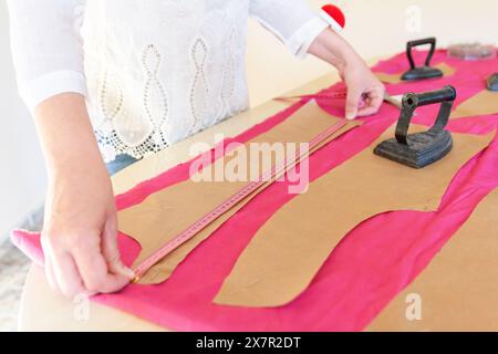 Close-up view of a tailor's hands using a measuring tape on pink and beige fabric, alongside tools like a vintage iron, highlighting precision in fash Stock Photo