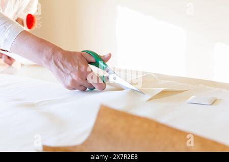 Close-up of a tailor’s hands cutting white fabric with green-handled scissors, with a pincushion on their wrist and brown paper patterns on the table Stock Photo