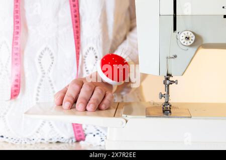 Close-up image showcasing a designer's hands operating a sewing machine with a pink measuring tape and a pincushion in the background suggestive of a Stock Photo