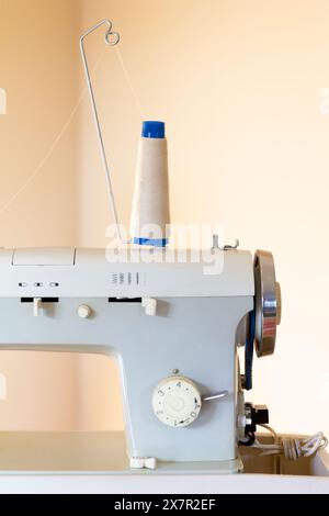 A close-up image of a professional sewing machine with a large spool of thread mounted on top, set against a beige background, representing a sewing a Stock Photo