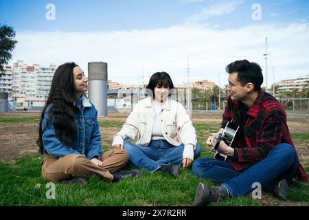 Three friends sharing a moment of joy in an urban park, with one playing the guitar. They look relaxed and happy. Stock Photo