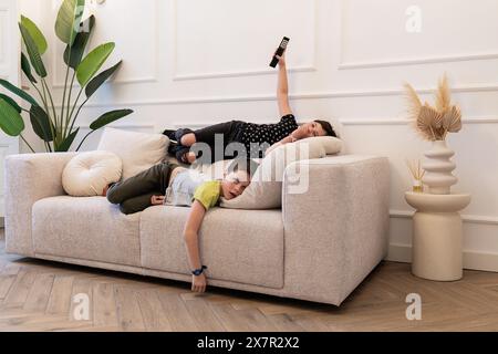 Two children lounging on a sofa at home, one lying on his stomach looking at the camera, the other stretching overhead with a remote, not looking at t Stock Photo