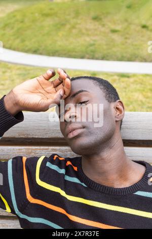 A young male, wearing a colorful striped sweater, sits on a park bench while appearing distressed and holding his head, indicating symptoms of hypogly Stock Photo