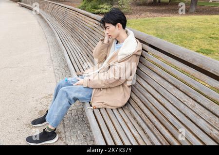 A young Asian male holds his head in discomfort while sitting on a park bench, possibly experiencing hypoglycemia symptoms Stock Photo