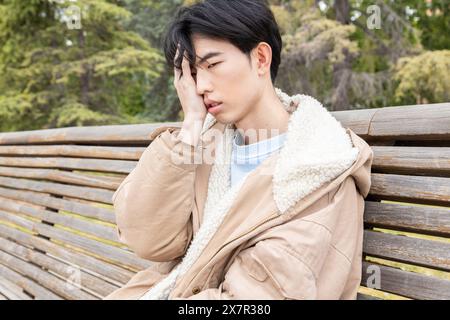 A young Asian man experiences symptoms of hypoglycemia while sitting on a park bench, looking distressed and holding his head Stock Photo