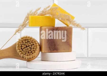A serene display of stacked handmade natural soaps, topped with delicate wheat spikes, next to a wooden body brush placed on a sleek, white surface Stock Photo