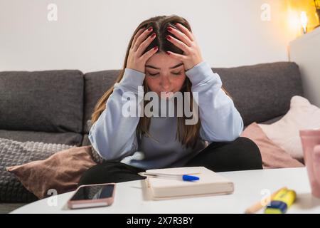A young woman seems overwhelmed and stressed, sitting on a couch with her hands on her head Stock Photo