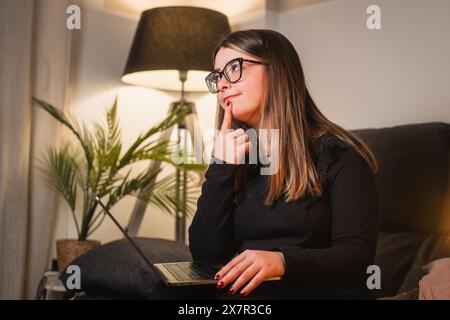A pensive young woman with glasses sits on a sofa at home, holding a laptop and touching her chin Stock Photo