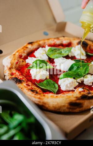 An anonymous person drizzles olive oil on a Margherita pizza topped with mozzarella and basil, freshly baked and served in a cardboard takeout box Stock Photo