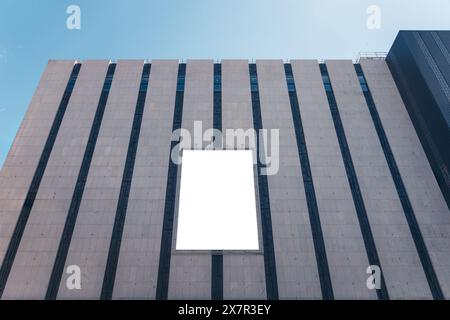 A mockup of a blank white billboard framed by a modern building facade under a clear blue sky in Madrid City, ideal for advertising Stock Photo