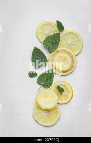 Top view of slices of bright yellow lemons arranged with green lemon balm leaves on a textured grey surface, ideal for culinary and natural themes. Stock Photo
