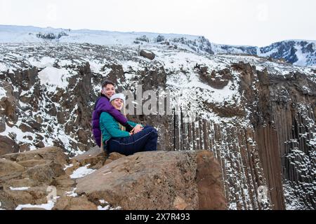 Young couple sitting on the snowy rocks of Iceland, smiling and posing against the dramatic backdrop of rugged volcanic cliffs partially covered with Stock Photo