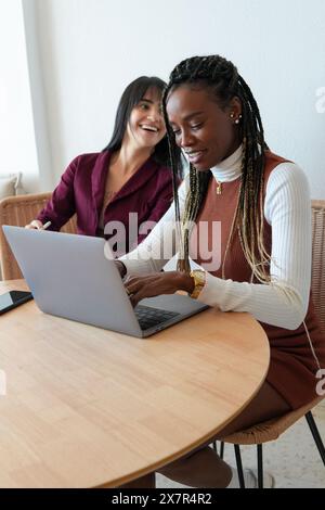 A cheerful scene of two diverse women working together on a laptop in a well-lit home environment, engaging in what appears to be a collaborative proj Stock Photo