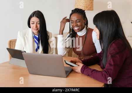 Three joyful women of diverse backgrounds sit at a table, engaging and collaborating on a laptop in a well-lit home setting They are clearly involved Stock Photo
