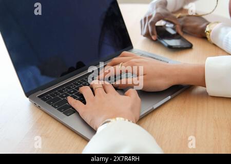 Close-up view of two women working together at a wooden desk, one typing on a laptop while the other uses a smartphone and mouse Their focus signifies Stock Photo