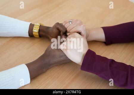 A close-up view capturing four people of diverse multiethnicities, joining hands together at a wooden table, symbolizing teamwork and unity Stock Photo